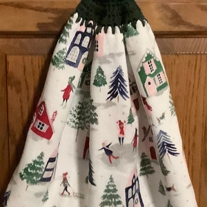 May include: A white kitchen towel with a winter scene print featuring houses, trees, and people. The towel has a green crocheted loop with a red button at the top.