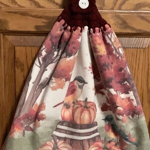 May include: A hanging kitchen towel with a fall theme. The towel features a watercolor design of pumpkins, birds, and autumn foliage in shades of orange, red, and brown. The top is a dark burgundy crochet with a white button.