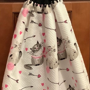 May include: White hanging kitchen towel with a black crocheted top and button closure. The towel features a repeating pattern of gray cats, pink hearts, and arrows. A ball of pink yarn is also in the design.