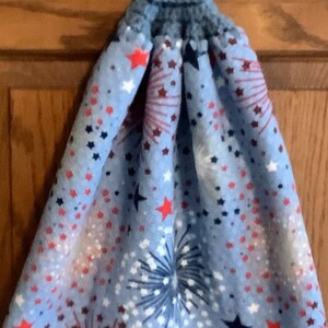 May include: Blue kitchen towel with a red, white, and blue firework and star pattern. The towel has a blue crocheted loop with a red button.