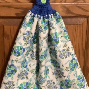 May include: A hanging kitchen towel with a blue crocheted top and a green button. The towel features a white background with a floral pattern in shades of blue and green. The towel is designed to hang from a hook.