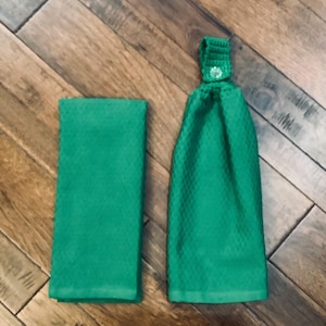 May include: Two green kitchen towels. One is folded, rectangular, and the other is a hanging towel with a crocheted top and a decorative button. The towels are made of a textured fabric.