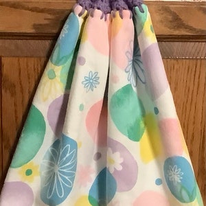 May include: A purple crocheted dish towel holder with a button. The towel is white with a colorful abstract floral pattern.
