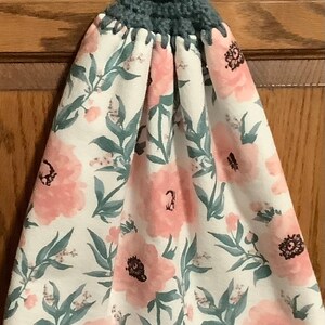 May include: A white kitchen towel with a floral pattern of pink flowers and green leaves. The towel has a gray crocheted loop with a button at the top.