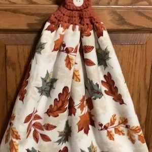 May include: A hanging kitchen towel with a fall leaf pattern. The towel features a white background with orange, brown, and green leaves. The top is crocheted in a rust-colored yarn and has a white button for hanging.