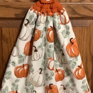 May include: A hanging kitchen towel with a white background featuring orange and white pumpkins and green leaves. The top portion is crocheted in orange yarn with a white button. A seasonal fall-themed kitchen accessory.
