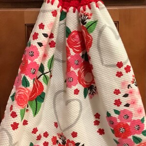 May include: A red crocheted kitchen towel topper with a white button. The towel features a white background with a floral pattern of pink and red roses, green leaves, and gray hearts. A decorative kitchen accessory.