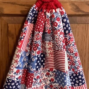 May include: A patriotic kitchen towel with a red crocheted top and button closure. The towel features a patchwork design with red, white, and blue patterns including stars, stripes, and floral elements. This decorative towel adds a festive touch to any kitchen.