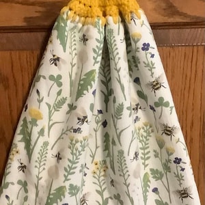 May include: A hanging kitchen towel with a yellow crocheted top and a green button. The towel features a white background with a floral and bee pattern in green, yellow, and purple.