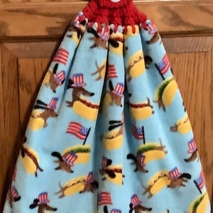May include: A hanging kitchen towel with a red crocheted top and a white button. The towel features a light blue background with a repeating pattern of hot dogs, dachshunds wearing patriotic hats, and American flags.