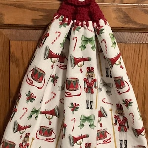 Puede incluir: Un porta-toallas de cocina de crochet rojo con un botón blanco. La toalla es blanca con un patrón repetido de cascanueces, tambores, trompetas, bastones de caramelo y lazos.