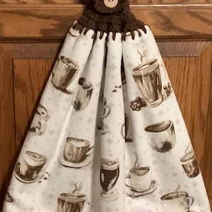 May include: A hanging kitchen towel with a brown crocheted top and button closure. The white fabric features a repeating pattern of coffee cups and coffee beans in shades of brown and gray.