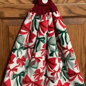 May include: A decorative kitchen towel with a red crocheted top and button closure. The towel features a white background with a pattern of red, green, and gray bows. The towel is designed for hanging.