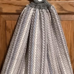 May include: A hanging kitchen towel featuring a textured, plush gray, white, and light blue striped pattern. The top section is made of solid gray crocheted fabric with a single white button closure, designed to hang securely from a cabinet handle.