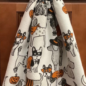 May include: A white kitchen towel with a Halloween theme featuring black and orange dogs wearing costumes. The towel has a black crocheted hanging loop with a black button.