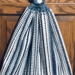 May include: A hanging kitchen towel with a blue and white pattern. The top portion is a solid blue crochet loop with a white button. The towel has a striped pattern in shades of blue and white, with a white striped border at the bottom.
