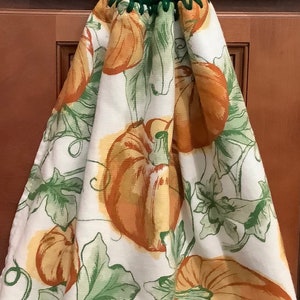 May include: A green crocheted dish towel holder with a brown button. The dish towel has a white background with a pattern of orange pumpkins and green leaves.