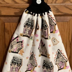 May include: White Halloween-themed kitchen towel with a black crocheted top and button closure. The towel features repeating illustrations of haunted houses, ghosts, and candy corn. The houses have pink accents and spiderwebs.