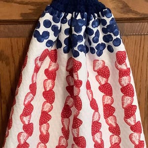 May include: A white kitchen towel with a red and white strawberry pattern and a blue crocheted hanging loop with a red button. The towel is folded in half and hanging on a wooden door.