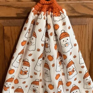 May include: A hanging kitchen towel featuring a white fabric with a repeating pattern of ghosts wearing orange knit hats, holding mugs or books, and surrounded by orange pumpkins and autumn leaves. The towel has an orange crocheted top with a black button.
