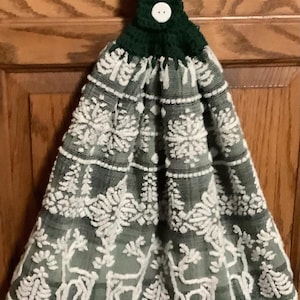 May include: A hanging kitchen towel featuring a green and white pattern of reindeer and snowflakes. The towel is topped with a dark green crocheted fabric loop secured by a single white button, allowing it to hang from a cabinet handle.