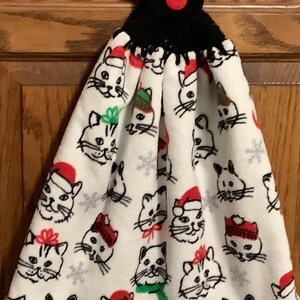 May include: White kitchen towel with a black crocheted loop and a red button. The towel features a repeating pattern of black and white cat faces wearing Santa hats and other festive accessories.
