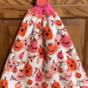 May include: A white hanging kitchen towel featuring a repeating pattern of orange and pink jack-o'-lantern pumpkins. The towel is topped with a pink crocheted fabric handle secured by a single orange button, designed for hanging on a cabinet handle.