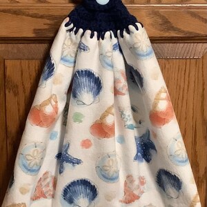 May include: A white kitchen towel with a blue and orange seashell pattern. The towel has a blue crocheted loop with a blue button.
