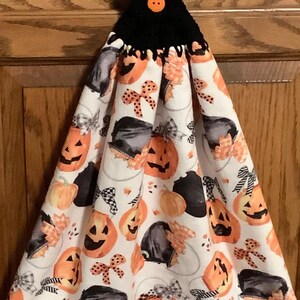 May include: A Halloween-themed kitchen towel with a black crocheted top and orange button. The white fabric features orange jack-o'-lanterns, black hats, and bows. A festive and decorative item for the fall season.