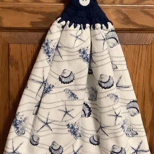 May include: A white kitchen towel with a blue and white seashell and starfish pattern. The towel has a navy blue crocheted loop with a white button.