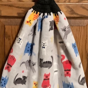 May include: A white kitchen towel with a black crocheted hanging loop and a button. The towel features a repeating pattern of colorful cartoon cats with different expressions.
