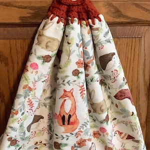 May include: A hanging kitchen towel with a crochet top and a button closure. The towel features a white background with a repeating pattern of woodland animals, including a fox, squirrel, and rabbit, surrounded by foliage.
