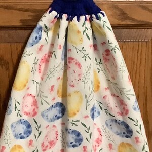 Puede incluir: Una toalla de cocina blanca con un patrón de huevo de acuarela azul, amarillo y rosa. La toalla tiene un lazo de crochet azul con un botón amarillo.