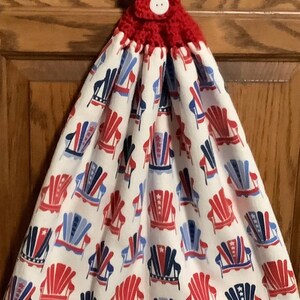May include: A red and white hanging kitchen towel with a button closure. The towel features a pattern of red, white, and blue Adirondack chairs, perfect for patriotic decor.