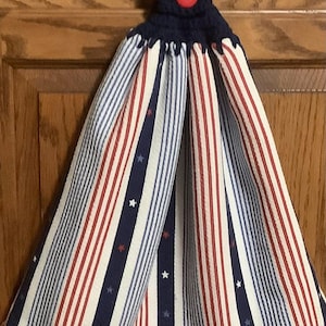 May include: A patriotic kitchen towel with a navy blue crocheted top and a red button. The towel features alternating red, white, and blue vertical stripes with small white stars, creating a festive design.
