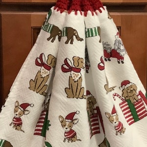 May include: A red crocheted dish towel holder with a white button. The towel features a white background with a repeating pattern of dogs wearing Santa hats and scarves.