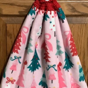 May include: A pink Christmas-themed hanging towel with a red crocheted top and button closure. The towel features a repeating pattern of Christmas trees in pink, green, and red, with bow accents. The towel is designed for kitchen or bathroom use.