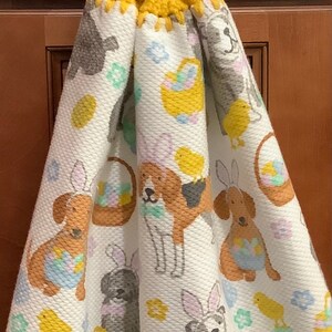 May include: A white kitchen towel with a yellow crocheted hanging loop. The towel features a repeating pattern of dogs wearing bunny ears and holding baskets of Easter eggs. There are also small yellow chicks on the towel.