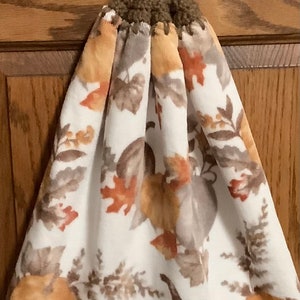May include: A white kitchen towel with a brown and orange pumpkin and leaf pattern. The towel has a brown crocheted loop with a brown button.