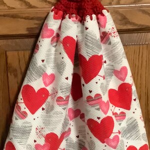 May include: A red crocheted dish towel holder with a white button. The towel is white with a red and pink heart pattern.