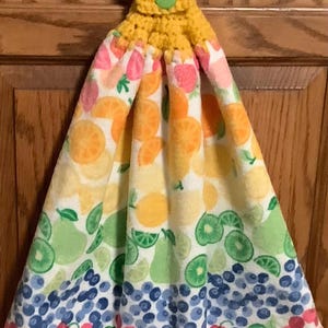 May include: A yellow crocheted kitchen towel with a green button closure. The towel features a colorful fruit pattern, including oranges, lemons, limes, blueberries, and cherries. The fruit design is printed on a white background.