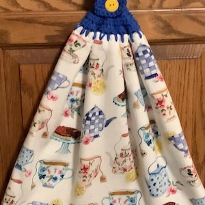 May include: A white kitchen towel with a blue crocheted top and yellow button. The towel features a pattern of teacups, teapots, and lemon slices with floral accents. The towel is designed to hang.