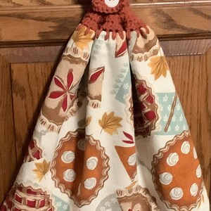 May include: A hanging kitchen towel featuring a white fabric with a repeating pattern of various pies, autumn leaves, and geometric shapes. The towel is secured at the top with a rust-colored crocheted tab and a single white circular button.