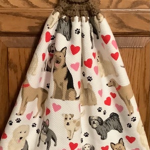 May include: A white kitchen towel with a brown crocheted hanging loop and a brown button. The towel features a pattern of dogs, hearts, and paw prints.