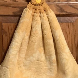 May include: A yellow kitchen towel with a sunflower design, hanging from a crocheted loop with a yellow button. The towel is made of a soft, absorbent material and has a white and yellow checkered border.