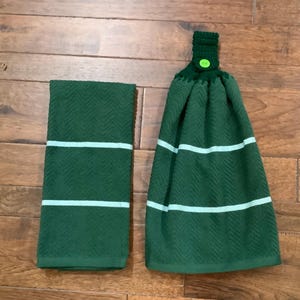 May include: Two-piece kitchen towel set. The set includes a dark green hand towel and a hanging towel. Both towels have a herringbone pattern with two light green stripes. The hanging towel has a crocheted top with a green button.