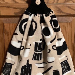 May include: A beige kitchen towel with black and white kitchen utensil designs. The towel has a black crocheted loop with a wooden button at the top.