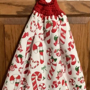May include: A hanging kitchen towel featuring a white fabric with a repeating pattern of red and white candy canes and wrapped candies. The towel is secured at the top by a red crocheted topper with a single white button for closure.