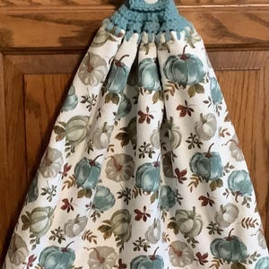 May include: A hanging kitchen towel featuring a white fabric base with a repeating pattern of teal and sage green pumpkins and brown leaves. The top section is made of teal crocheted yarn with a single white button for securing the towel.