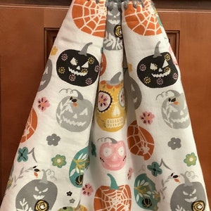 May include: A white kitchen towel with a Halloween theme featuring various jack-o'-lantern designs in orange, black, gray, and pink. The towel has a gray crocheted loop with an orange button.
